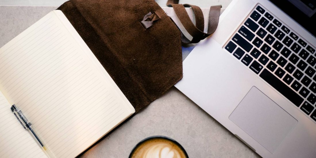 An open notebook and pen, leather satchel, laptop and coffee on a desk