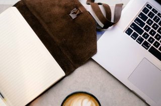 An open notebook and pen, leather satchel, laptop and coffee on a desk
