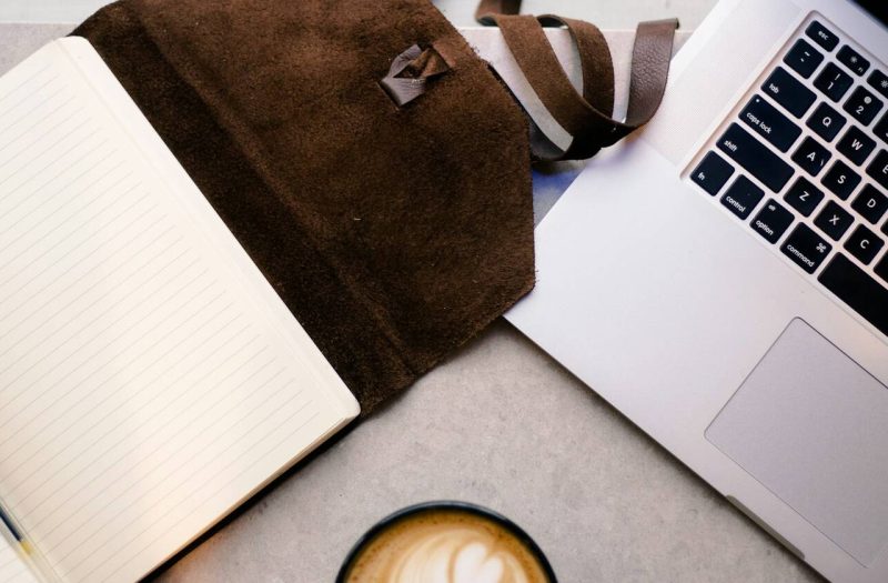 An open notebook and pen, leather satchel, laptop and coffee on a desk