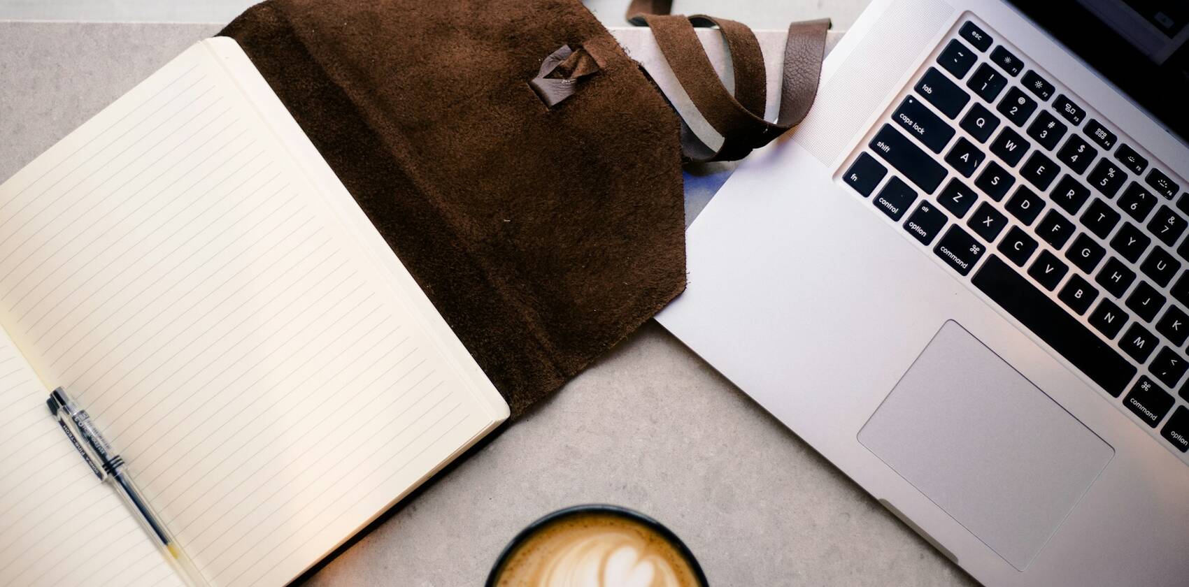 An open notebook and pen, leather satchel, laptop and coffee on a desk